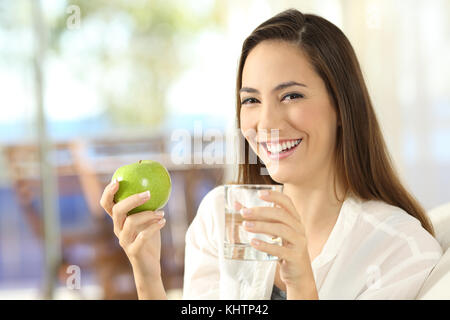 Donna felice tiene una mela e un bicchiere di acqua seduto su un divano nel soggiorno di casa Foto Stock