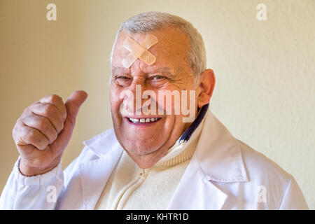 Il vecchio medico con patch sulla fronte sorridente e mostrando pollice in alto Foto Stock