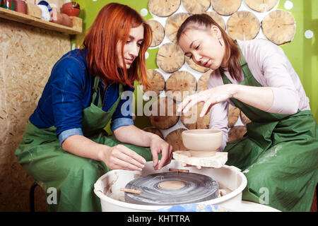 Una giovane donna potter in un grembiule verde insegna a scolpire una donna su un tornio del vasaio un vaso fatto di argilla marrone, racconta e mostra come lavora, vie laterali Foto Stock