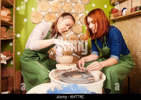 Una giovane donna potter in un grembiule verde insegna a scolpire una donna su un tornio del vasaio un vaso fatto di argilla marrone, racconta e mostra come lavora, vie laterali Foto Stock