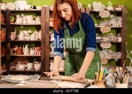 Una giovane donna potter in un grembiule verde rotola fuori una gabbia di creta per fare una piastra, su di un tavolo di legno ci sono strumenti e spazzole in background, rack Foto Stock