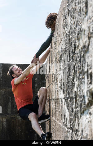 Giovane maschio free climber alla sommità della parete del mare aiutando un amico di salire Foto Stock