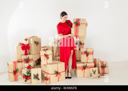 Giovane donna in abito rosso per impilata regali di Natale tenendo il pacco di Natale Foto Stock