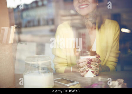 Giovane donna seduta al cafe, tenendo tazza da caffè, tatuaggi sul lato vista attraverso la finestra cafe, metà sezione Foto Stock