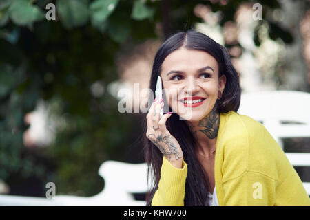 Giovane donna seduti all'aperto, utilizza lo smartphone, sorridente, tatuaggi a portata di mano e collo Foto Stock