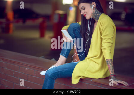 Giovane donna tenendo lo smartphone, indossa gli auricolari, sorridente, tatuaggi sul braccio a mano a mano e collo Foto Stock