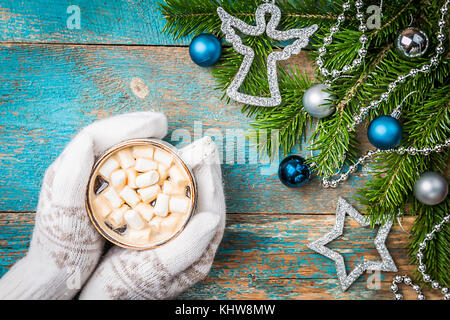Le mani delle donne in muffole tenendo una tazza di una bevanda calda su un blu tavolo in legno vista superiore. decorazioni di Natale e una tazza di cioccolata calda. Foto Stock