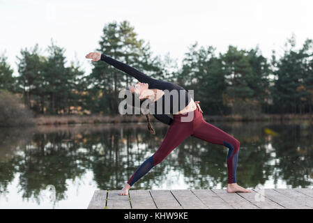 Donna pratica lo yoga da un lago facendo un guerriero retromarcia pongono. Foto Stock