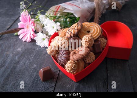 Biglietto di auguri idea con una a forma di cuore scatola piena di gustosi biscotti, cioccolatini e un mazzo di fiori avvolti in un giornale in background. Foto Stock