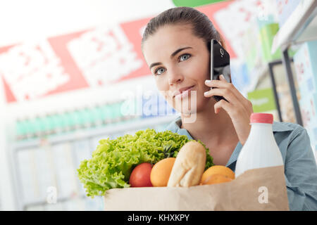 Bella giovane donna fare la spesa al supermercato, sta avendo una telefonata e tenendo un sacchetto con verdure fresche Foto Stock