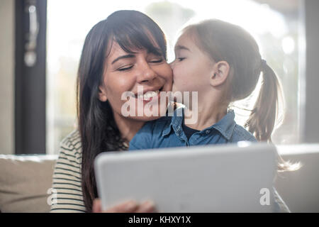 Donna baciato sulla guancia dalla figlia sul divano mentre usando tavoletta digitale Foto Stock