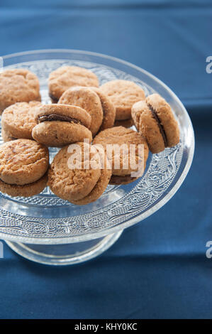 Lady's baci (pasticcini di pasta di mandorle e cioccolato) sulla torta di vetro si erge lavorato. blu di prussia tovaglia come sfondo. Foto Stock