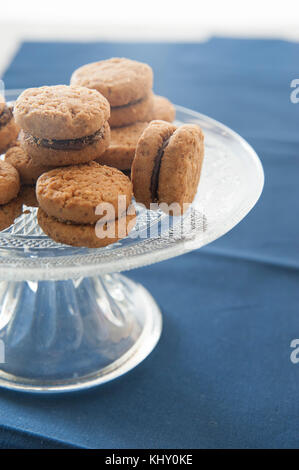 Lady's baci (pasticcini di pasta di mandorle e cioccolato) sulla torta di vetro si erge lavorato. blu di prussia tovaglia come sfondo. Foto Stock