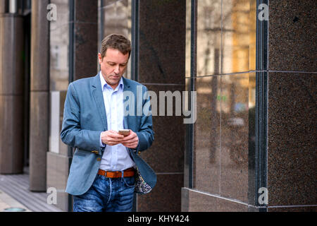 Un uomo d'affari di mezza età che cammina accanto all'edificio dell'ufficio mentre usa il suo smartphone Foto Stock