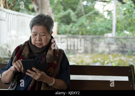 Il sambuco donna tenendo il telefono cellulare seduta sul banco in giardino. anziani femmina messaggio sms utilizzando app con il cellulare nel parco. senior utilizza lo smartphone f Foto Stock