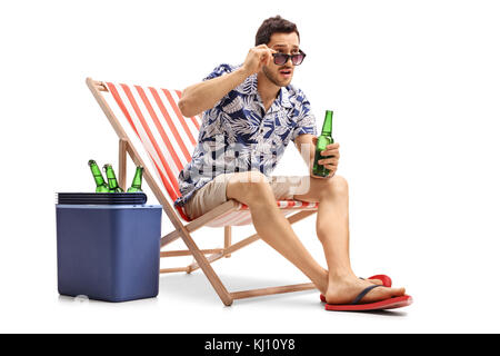 Stupito turistico con una bottiglia di birra seduto in una sedia a sdraio e fissando sopra i suoi occhiali da sole isolato su sfondo bianco Foto Stock