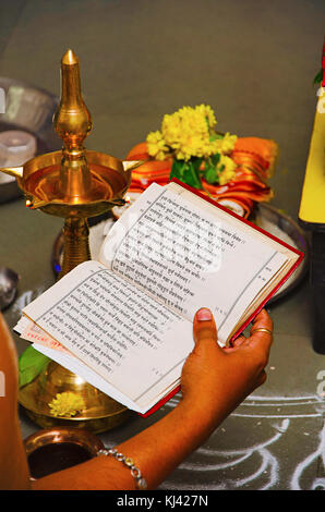 Sacerdote tenendo un libro contenente i rituali nella sua mano destra con una tradizionale lampada, fiori e rangoli in background, Belgavi, Karnataka, India Foto Stock