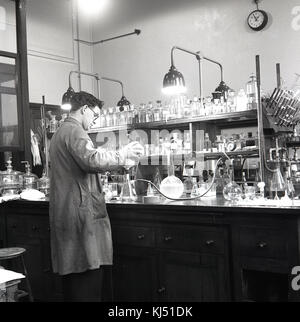 Degli anni Cinquanta, storico ricerca maschio scienziato che lavora in un laboratorio chimiche con numerose bottiglie di vetro e contenitori su un banco di lavoro. Foto Stock