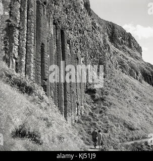 Degli anni Cinquanta, storico, un paio di camminare insieme lungo uno stretto sentiero costiero accanto a una ripida scogliera che mostra le antiche basalto vulcanico colonne e formazioni rocciose che rendono il Selciato del gigante in Co Antrim, Irlanda del Nord, un luogo così speciale da visitare. Foto Stock