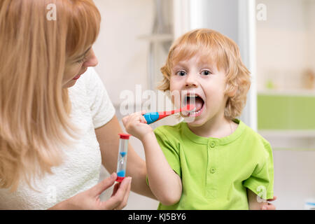 Happy Child boy spazzolare i denti vicino a specchio in bagno. sua madre Monitoraggio della precisione e tempo di azione di pulizia con la clessidra. Foto Stock