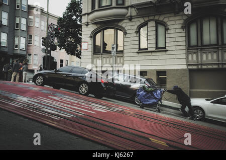 Uomo senza tetto spingendo un carrello in salita su una ripida collina di San Francisco, California Foto Stock