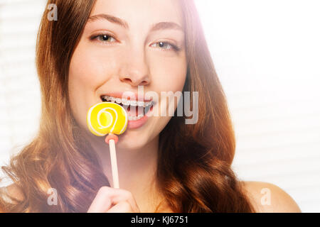 Ritratto di bella dai capelli scuri donna con bretelle dentali mordere off swirl giallo lecca-lecca, contro sfondo tranciato Foto Stock