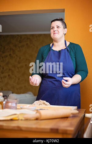 Giovane bella donna con capelli corti, in piedi in cucina dietro un tavolo da cucina, sorridente guardando verso l'alto. le sue mani sono coperte con la farina, ha preso Foto Stock