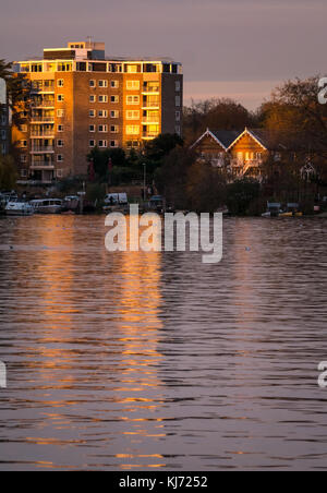 In autunno i colori del tramonto sul fiume Tamigi, Hampton Wick guardando verso est lungo il fiume ad un alto e moderno blocco di appartamenti riflessi nell'acqua, London, Regno Unito Foto Stock