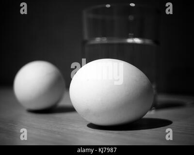 Foto in bianco e nero di due uova e un bicchiere di acqua su un sfondo di parete Foto Stock