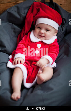 Baby girl in santa claus costume e hat siede a piedi nudi tra cuscino e sorrisi. Foto Stock