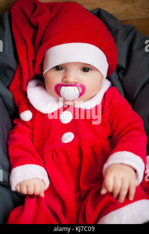 Baby girl in santa claus costume e hat siede tra cuscino con il succhietto nella bocca. primo piano. Foto Stock