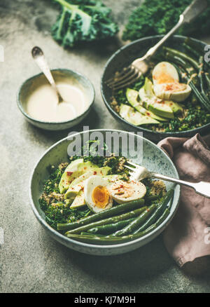 Una sana colazione vegetariana bocce. La quinoa, kale, fagioli verdi, avocado, uovo e cremoso condimento tahini bocce su grigio sfondo concreto, selettivo Foto Stock