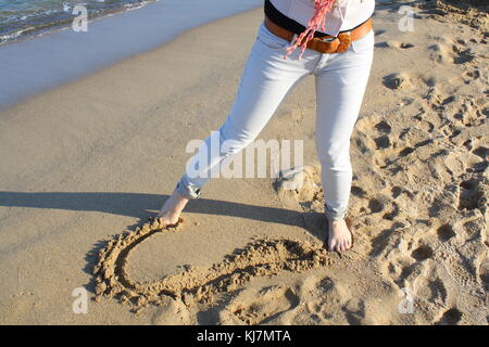 Una donna richiama un cuore nella sabbia sulla spiaggia per mostrare il concetto del suo amore sentimenti. Foto Stock