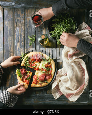 Tenendo le mani a fette di pizza fatta in casa con formaggio e bresaola, servita su piastra nera con rucola fresca, olio di oliva, un bicchiere di vino rosso e asciugamano da cucina Foto Stock