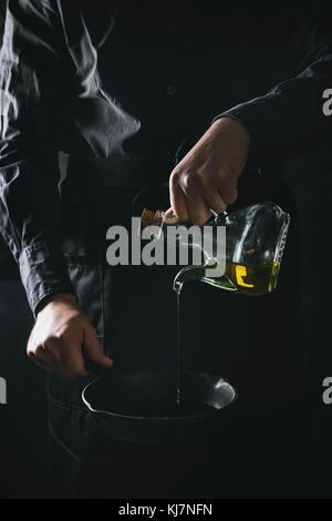 Lo chef uomo in nero grembiule versando olio d'oliva dalla bottiglia per cucinare frittelle in ghisa pan. Scuro in stile rustico. Immagine dai toni Foto Stock