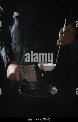 Lo chef uomo in nero grembiule versando l'impasto dalla siviera per cucinare frittelle in ghisa pan. Scuro in stile rustico. Foto Stock