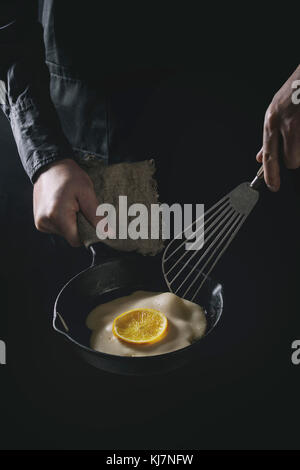 Lo chef uomo in nero grembiule cucinare frittelle con orange in ghisa pan. Trasforma i pancake. Scuro in stile rustico. Immagine dai toni Foto Stock