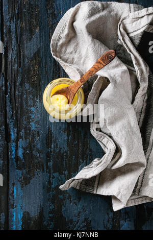 Bicchiere di tratte di ghee burro in piedi con il cucchiaio di legno e tessuto di lino sopra il vecchio colore blu scuro dello sfondo di legno. Mangiare sano. Vista superiore, spazio di copia Foto Stock