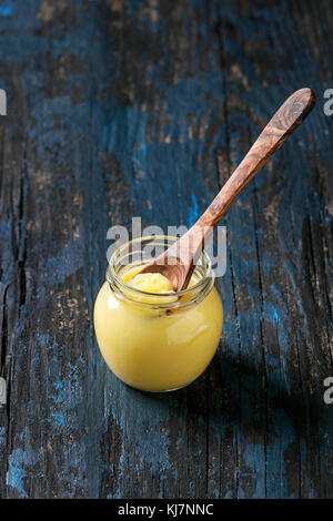 Bicchiere di tratte di ghee burro in piedi con il cucchiaio di legno sopra il vecchio colore blu scuro dello sfondo di legno. Mangiare sano. Foto Stock