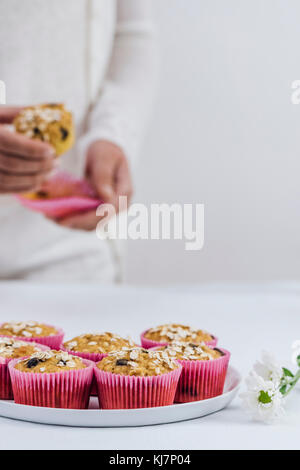 Una donna in bianche vesti la rimozione di un muffin di zucca da una rosa di muffin liner fotografata da vista frontale. Una piastra piena di muffin e fiori bianchi su Foto Stock
