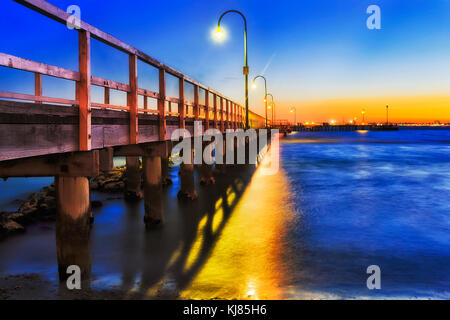 Dark tramonto sul centro storico pontile in legno a Port Melbourne sulla spiaggia della Baia di Port Phillip. Le luci di strada e il sole di setting riflettono in sfocata acque blu. Foto Stock