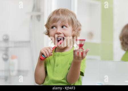 Bambino felice spazzolare i denti vicino a specchio in bagno. Egli è il monitoraggio della durata di azione di pulizia con la clessidra. Foto Stock