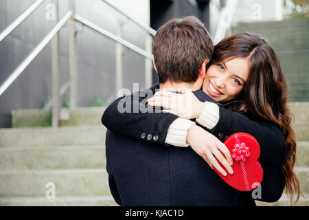 Bella e felice giovane donna in amore abbracciando il suo fidanzato tenendo un cuore rosso a forma di scatola regalo Foto Stock