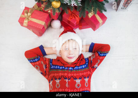 Closeup ritratto di sorridere grazioso fanciullo in vacanza di Natale vicino interno albero festoso. vista dall'alto del capretto felice posa su bianco pavimento in legno vicino Foto Stock