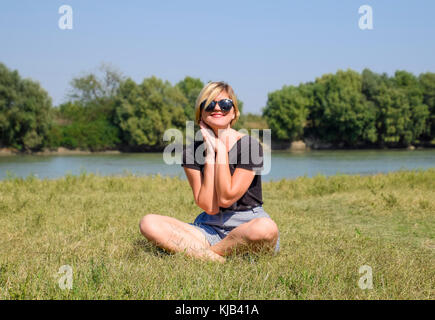 Ragazza con occhiali da sole si siede sul prato di una Lotus pongono. il biondino di sorrisi e mette le sue palme alla sua guancia. ragazza sullo sfondo del fiume Foto Stock