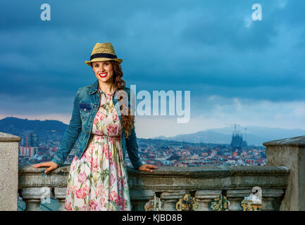 Serata perfetta con vista mozzafiato. ritratto di sorridente turistica elegante donna in abiti lunghi e cappello di paglia contro il paesaggio urbano di Barcellona, Spagna Foto Stock