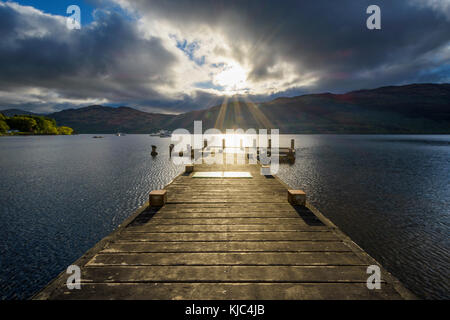 Molo di legno sul lago con le nuvole drammatiche all'alba a Loch Lomond in Scozia, Regno Unito Foto Stock