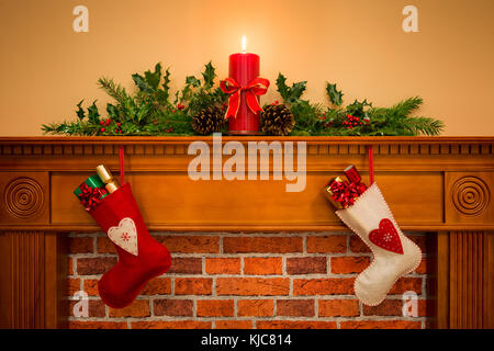 Due calze di Natale con regalo presenta appeso su un mantelpiece oltre un caminetto, plus candela che brucia con festosa ghirlanda compresi holly Foto Stock