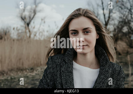 Bella e giovane donna con capelli lunghi biondi, ampia sopracciglia, tutte le labbra, occhi blu vestiti di lana cappotto. Ragazza guardando la fotocamera. Giunco di palude di sfondo. Foto Stock