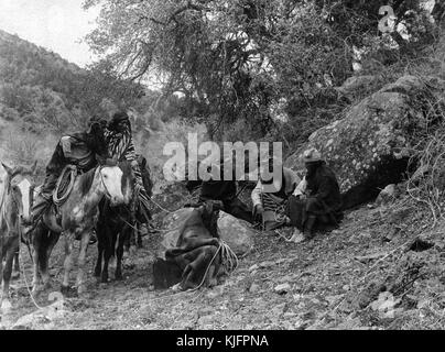 Fotografia di un gruppo di nativi americani, gli uomini seduti per terra nel deserto, i loro cavalli in piedi a sinistra, intitolato 'apache, la narrazione di gruppo di White Mountain apache', da Edward s Curtis, 1904. dalla biblioteca pubblica di new york. Foto Stock